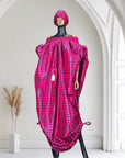 Pink off-shoulder boubou dress with black grid pattern, paired with a matching headwrap, displayed on a mannequin against a black paneled wall.
