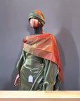 Olive green satin gown with ruched side draping, paired with a bronze-toned shawl and matching headwrap, displayed on a mannequin in an arched interior.
