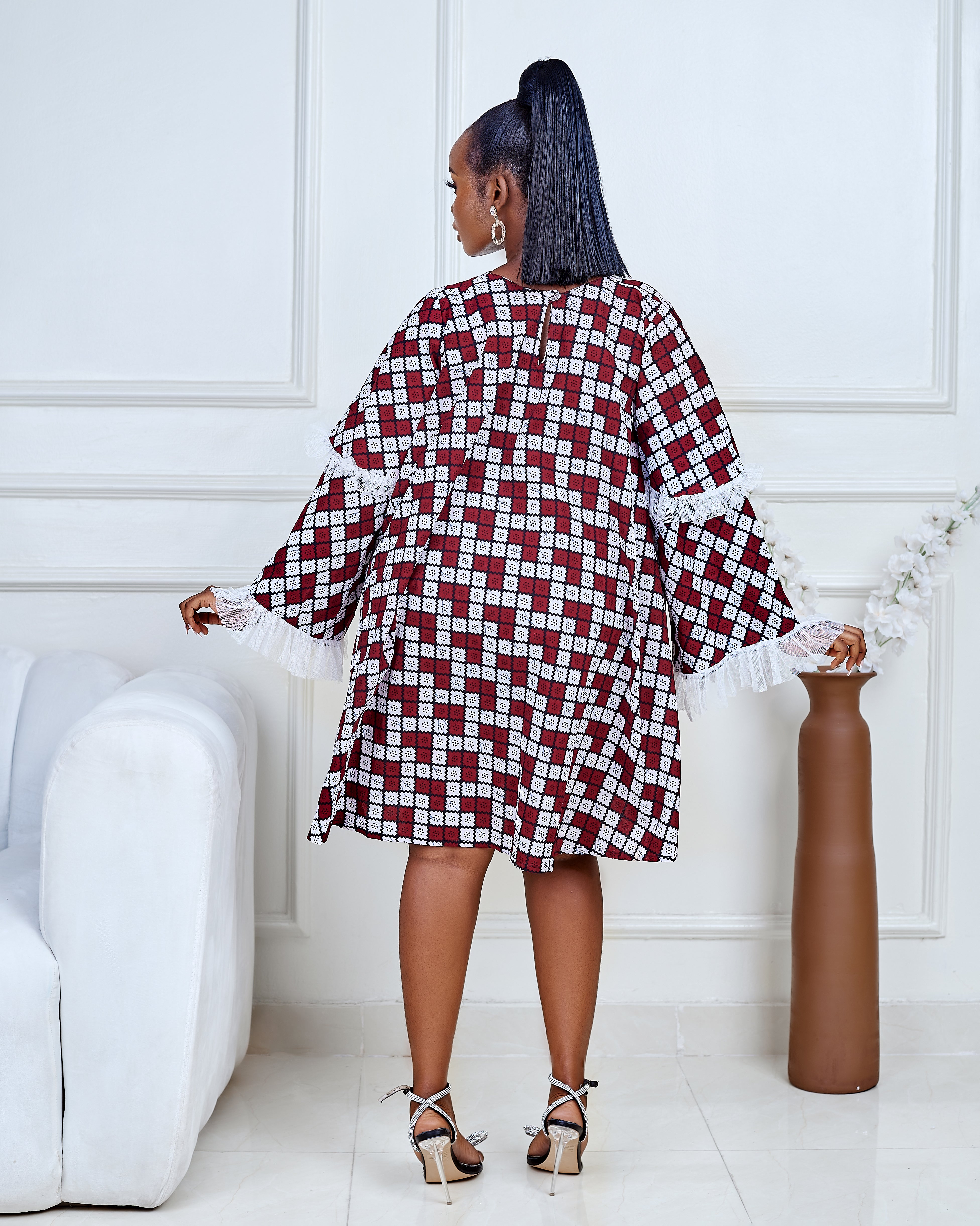 Model wearing a short flared Ankara dress with white lace sleeve detailing in a minimalist living room setting.
